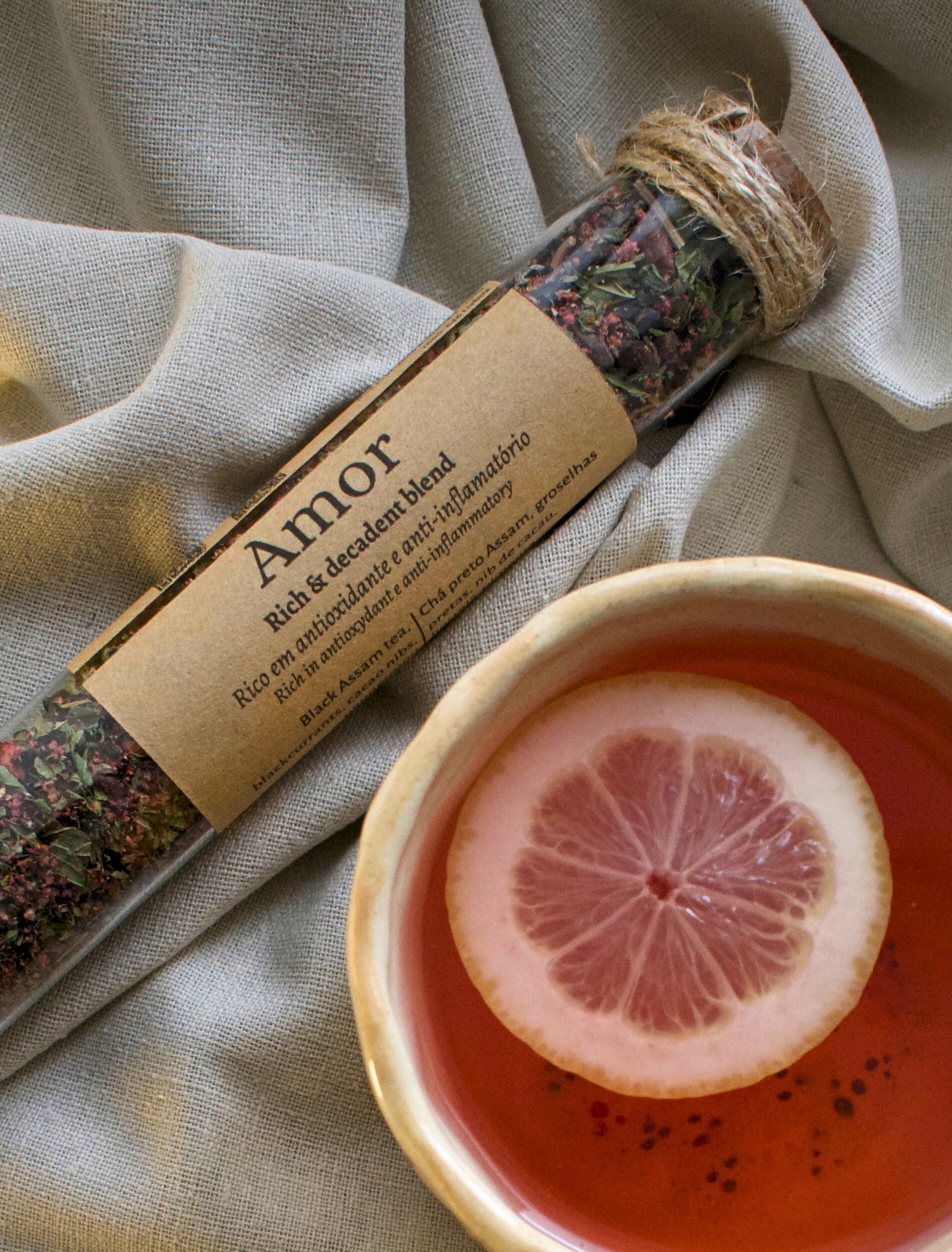 Amor tea blend in a glass tubes, next to a tea cup with a slice of lemon on a linen cloth.