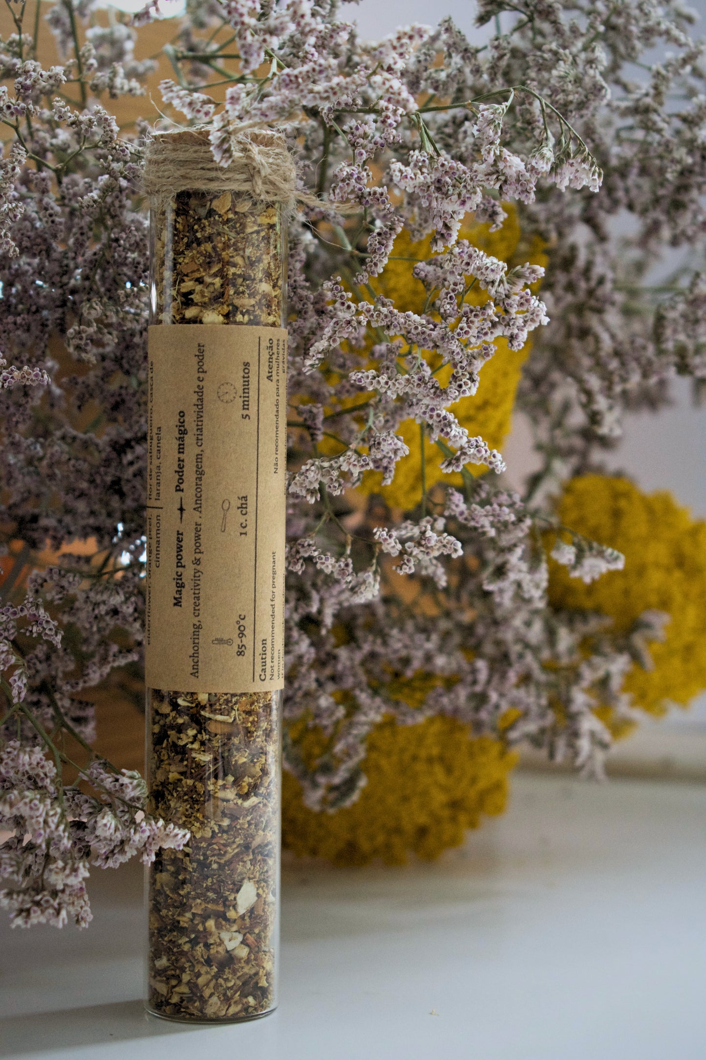 Dried herbs in a glass tube with a label, surrounded by flowers on a white surface