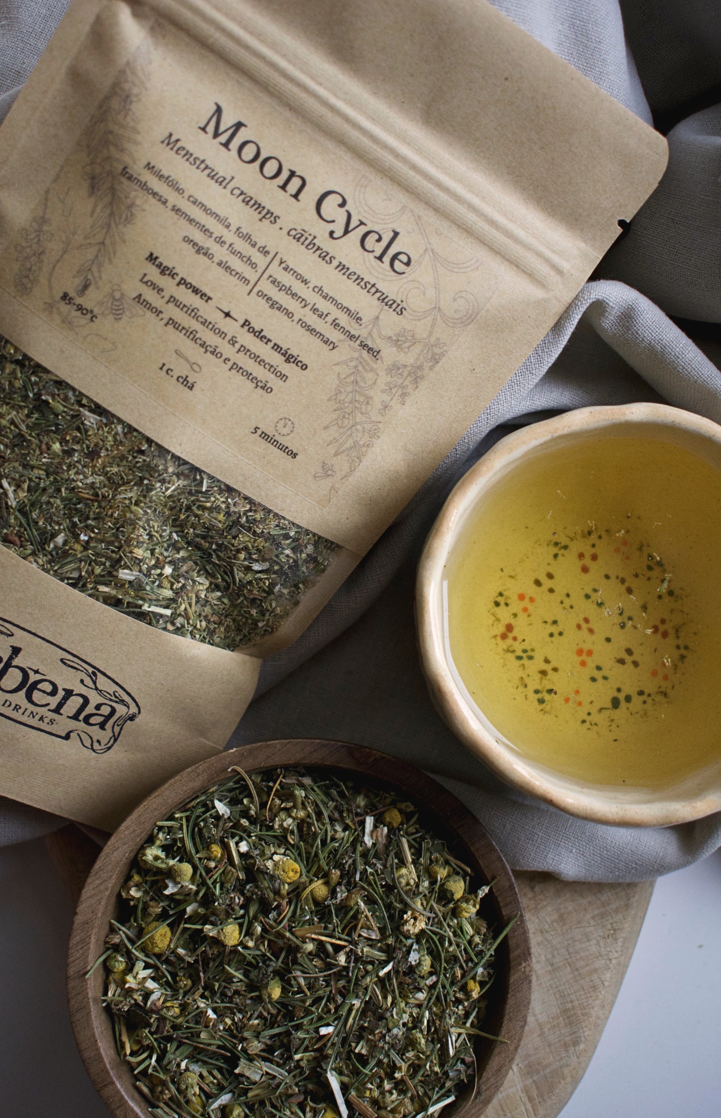 Moon Cycle kraft bag with a tea cup and the herbal blend in a wooden bowl on a linen cloth background.
