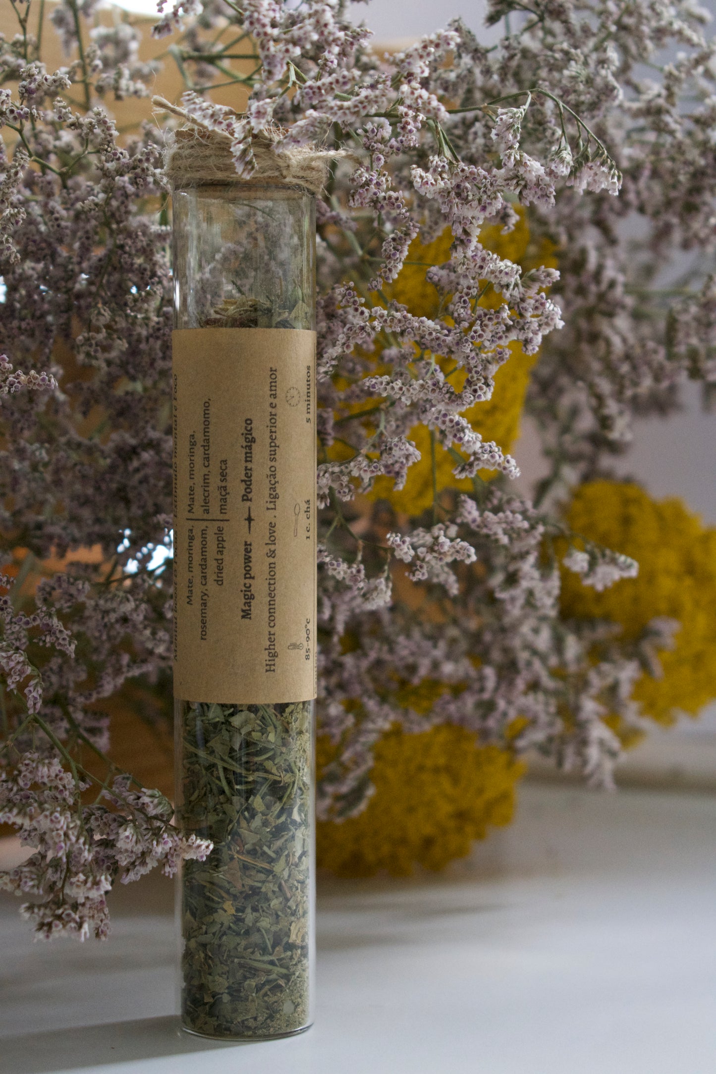 Glass tube with dried herbs on a white surface with flowers in the background