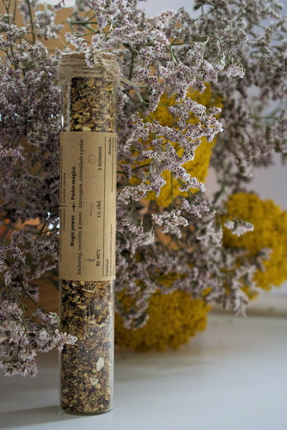 Dried herbs in a glass tube with a label, surrounded by flowers on a white surface